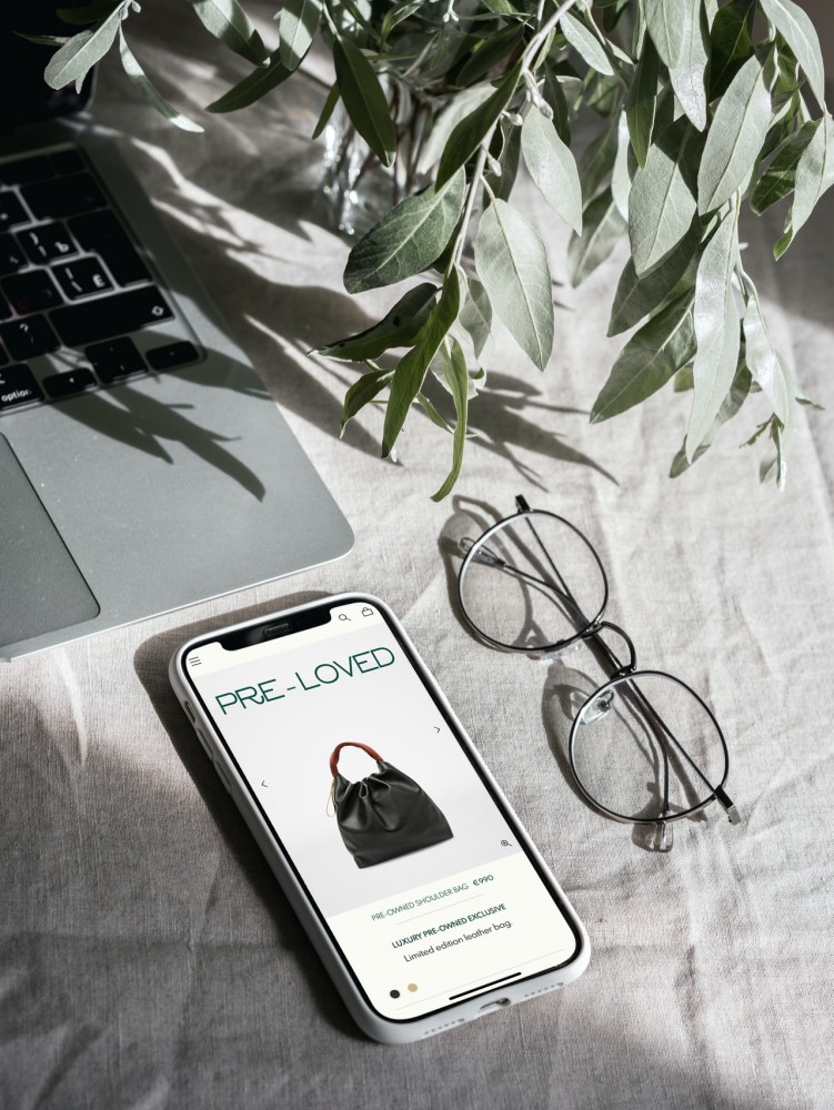 A smartphone that shows the page of an online luxury store that displays a handbag. On the image it says 'pre-loved' above the handbag.