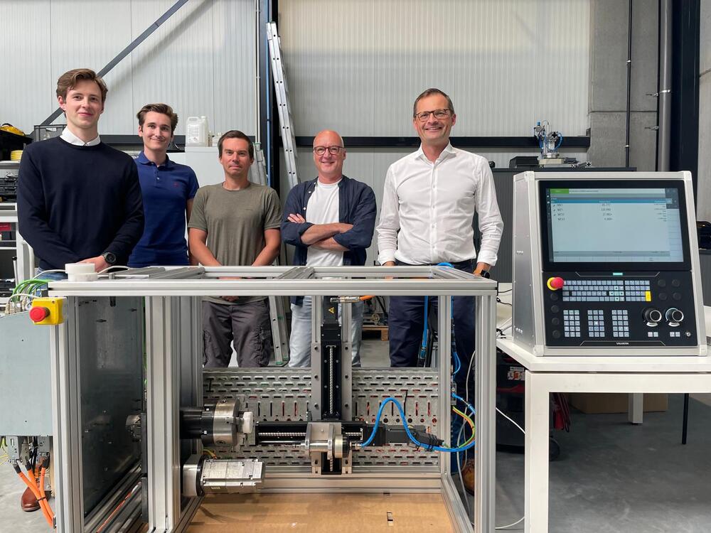 Foto tijdens de proefverdediging van deze bachelorproef, met onderaan het prototype en rechts de Siemens Sinumerik-controller. Van rechts naar links: de managing director van Cadcamatic, de externe mentor, de interne mentor, de mechanisch ingenieur en de student
